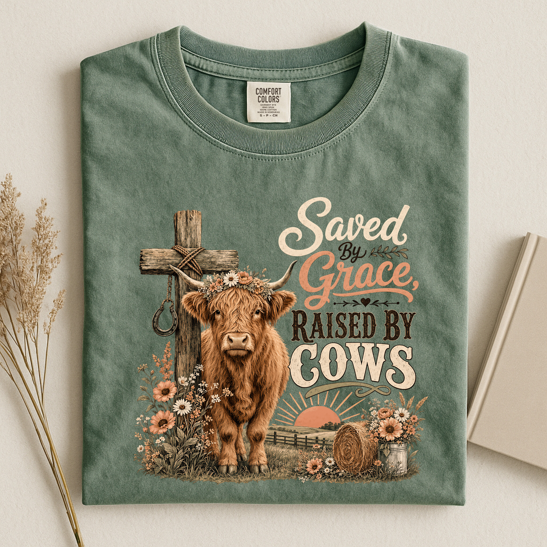A folded sage green t-shirt with a rustic Christian cow graphic, displayed on a light beige background with dried grass and a book as props. The graphic features a highland cow, a wooden cross, and the text 'Saved By Grace, Raised By Cows'.