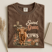 A brown short-sleeve t-shirt with a rustic graphic print of a highland cow, a cross, and the text 'Saved by Grace, Raised by Cows'. The shirt is displayed flat on a light beige background with dried grass on the left and a book on the right.