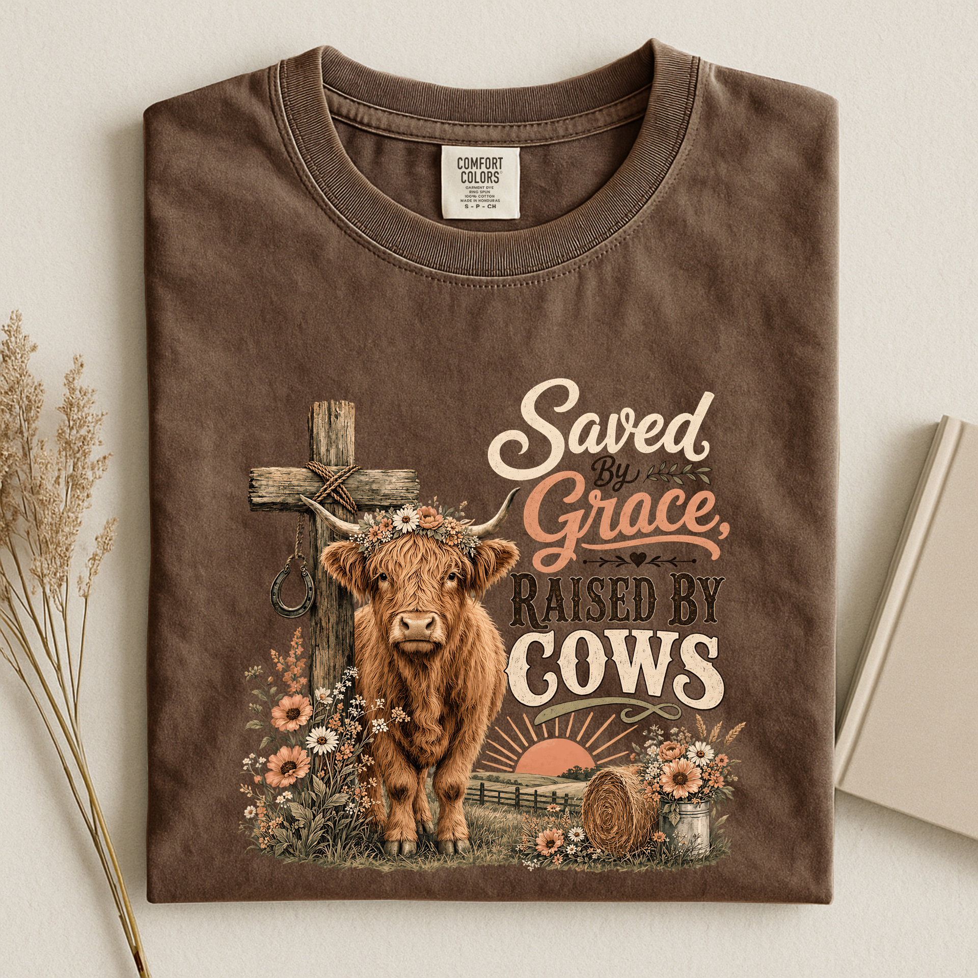 A brown short-sleeve t-shirt with a rustic graphic print of a highland cow, a cross, and the text 'Saved by Grace, Raised by Cows'. The shirt is displayed flat on a light beige background with dried grass on the left and a book on the right.
