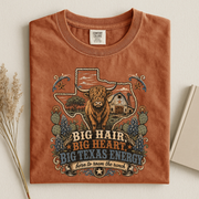 A folded terracotta-colored t-shirt with a detailed graphic print of a highland cow, Texas state outline, cactus, and a barn, with the text 'BIG HAIR, BIG HEART, BIG TEXAS ENERGY' and 'born to roam the ranch'.