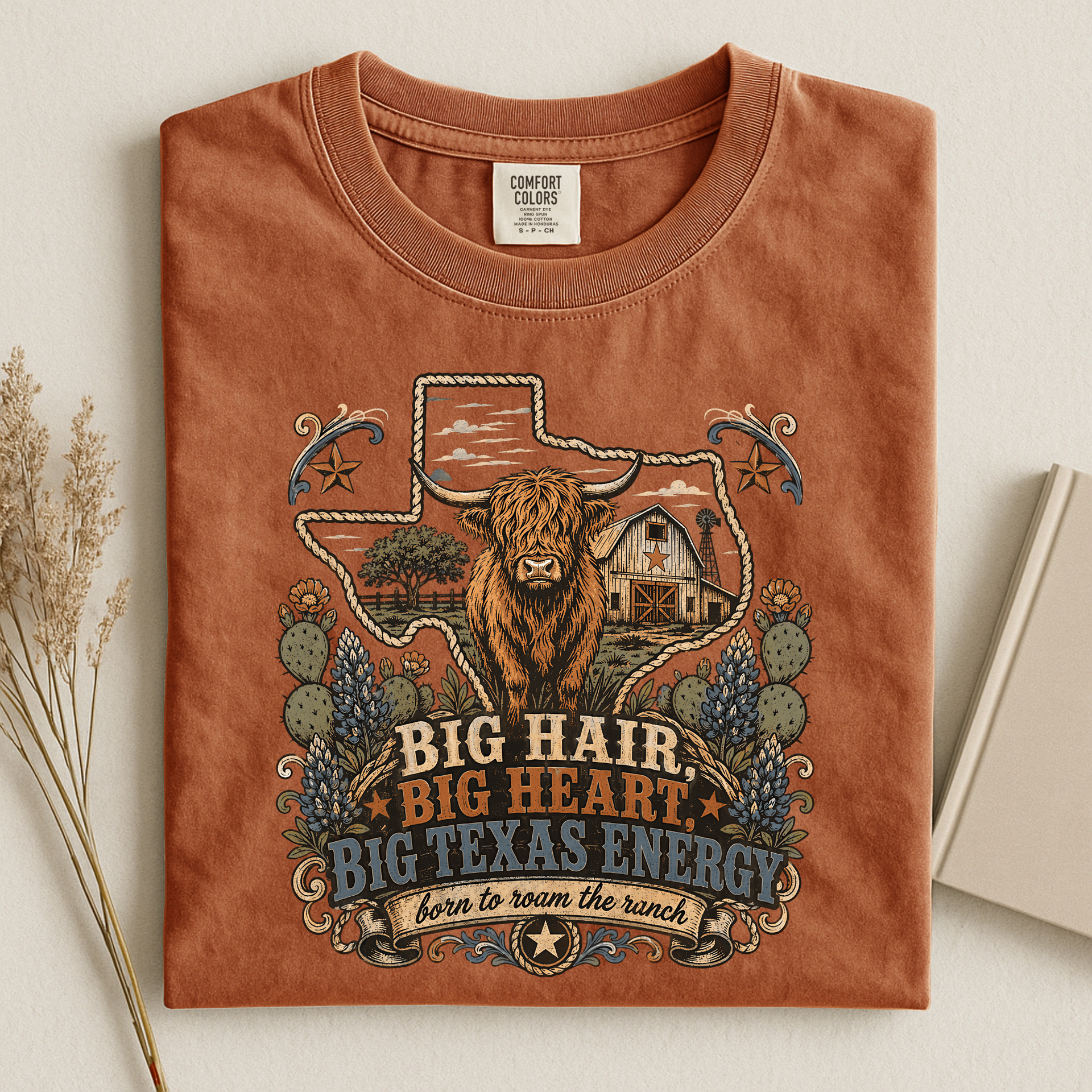 A folded terracotta-colored t-shirt with a detailed graphic print of a highland cow, Texas state outline, cactus, and a barn, with the text 'BIG HAIR, BIG HEART, BIG TEXAS ENERGY' and 'born to roam the ranch'.