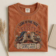 A rust-orange folded graphic T-shirt displayed on a light beige background with dried flowers and a book. The shirt features a western-themed graphic with cowboy boots, an American flag, a steer, and text reading 'Land of the Free Home Feisty' and 'cowgirl born & barn raised.